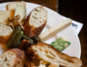 Cheese plate appetizer with crusty baguette slices, assorted cheeses including a green-marbled wedge and a firm white stick, cornichons and walnuts on a paper plate.
