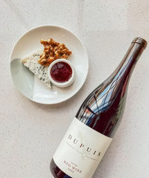 Red wine bottle lying next to a small plate with blue cheese wedge, candied walnuts, and red fruit jam on a light speckled countertop — a playful wine-and-cheese pairing.