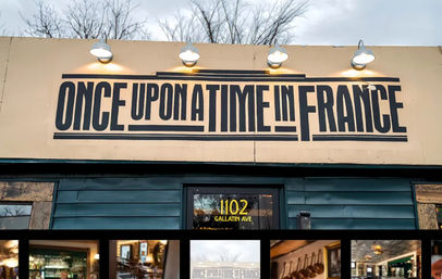 Teal street storefront with a large retro black-and-cream sign above the entrance, four metal lamps casting warm light, bare winter trees behind the building, and a row of small photos visible in the lower window.