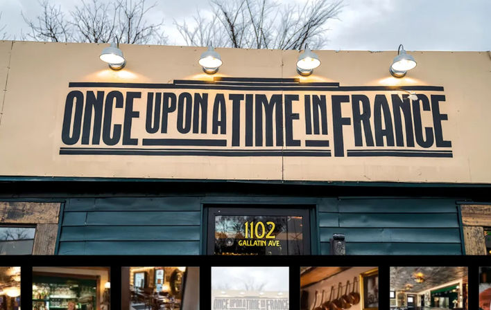 Teal street storefront with a large retro black-and-cream sign above the entrance, four metal lamps casting warm light, bare winter trees behind the building, and a row of small photos visible in the lower window.