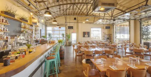 Bright modern industrial restaurant interior with a long wooden bar and turquoise metal stools, open dining area of wooden tables and chairs, exposed-beam ceiling, pendant lights and large windows flooding the space with natural light.