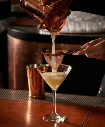 Bartender pouring a silky pale-yellow cocktail through a copper fine-mesh strainer into a martini glass on a leather-topped bar counter with a copper shaker nearby.