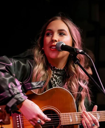 Close-up of a singer-songwriter with pink-tinted wavy hair, wearing a leather jacket and leopard-print top, singing into a microphone and playing an acoustic guitar on a live indoor stage