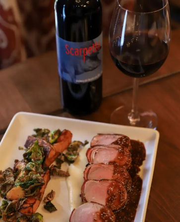 Gourmet restaurant plate of sliced pork tenderloin with mustard sauce, roasted sweet potatoes and Brussels sprouts, accompanied by a glass and bottle of red wine on a wooden table