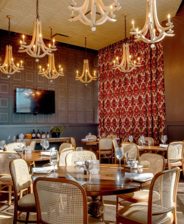 Elegant restaurant interior with glowing white chandeliers, round wooden tables, woven rattan chairs, red patterned drapery and neatly set wine glasses