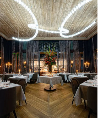 Elegant fine-dining restaurant interior with a looping crystal chandelier, candlelit tables, central floral centerpiece and chevron wood floor.