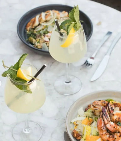 Two lemon-mint cocktails with lemon wedges and metal straws beside a grilled shrimp salad on a marble table, bright summer brunch scene.