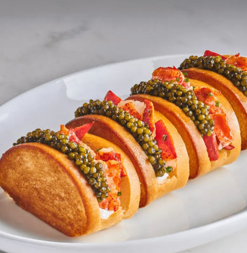Fine-dining seafood appetizer: row of lobster-and-caviar mini blini bites with tender lobster meat and glossy caviar on golden mini pancakes, plated on a white dish.