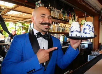 Smiling bartender in a bright blue tuxedo and handlebar mustache pointing to a tray with two blue-and-white ceramic cocktail cups garnished with lime at a stocked bar