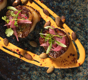 Two seared scallops on a speckled blue plate with a swipe of spicy orange aioli, topped with pickled red onions, microgreens and pumpkin seeds — elegant seafood appetizer