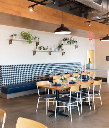 Bright modern urban café dining area with wooden tables and blue-seat chairs, houndstooth-pattern banquette, white wall shelves holding potted plants, pendant lights and exposed ductwork — inviting brunch-ready seating.