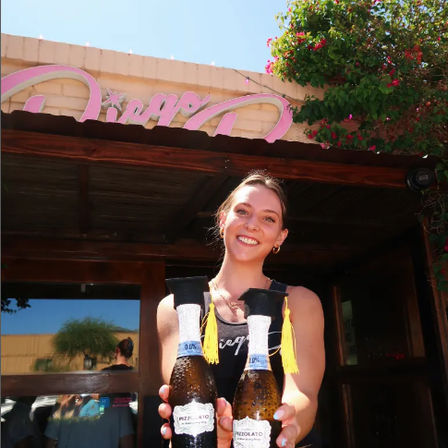 Smiling server holding two sparkling-wine bottles topped with tiny decorative caps, standing outside a sunny cafe patio under a pink sign with bougainvillea accents.