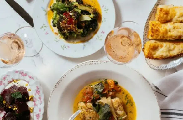Top-down view of a Mediterranean-style meal on a white tablecloth: bowls of vegetable stew with herbs, beet salad, crusty bread slices and glasses of rosé wine on vintage floral plates.
