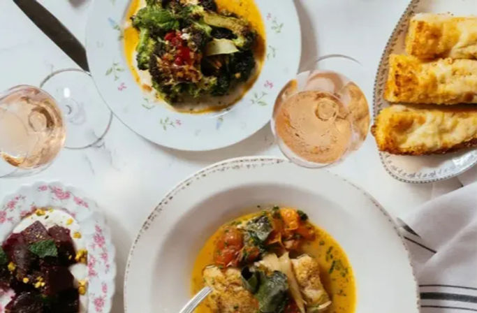 Top-down view of a Mediterranean-style meal on a white tablecloth: bowls of vegetable stew with herbs, beet salad, crusty bread slices and glasses of rosé wine on vintage floral plates.