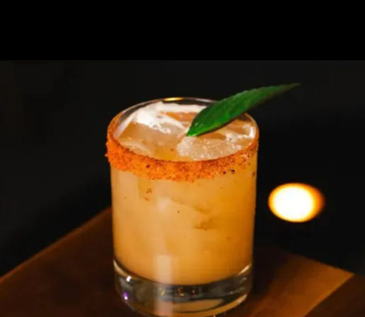 Zesty orange cocktail in a lowball glass with ice, chili-spiced rim and a green leaf garnish on a dim bar background