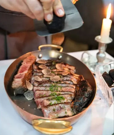 Candlelit, decadent sliced ribeye in a copper pan topped with shaved black truffle as a chef grates more — elegant restaurant plating