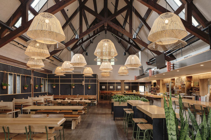 Airy rustic cafe interior with vaulted timber beams and skylights, oversized woven pendant lights, long communal wooden tables, bar seating and indoor plants.