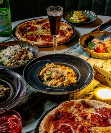 Italian dinner spread on a marble table in a cozy bistro: pepperoni pizzas, creamy grated-cheese pasta, plated salads, sparkling water bottle and a wine glass