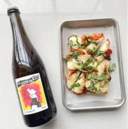 Top-down view of a dark wine bottle with a colorful illustrated label paired with a rectangular cheesy tomato-basil flatbread on a metal tray, arranged on a white speckled countertop.