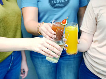 Three people toasting at a casual brunch — glasses of orange juice, a garnished cocktail, and sparkling wine.