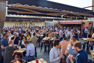 Busy urban rooftop bar patio at sunset with string lights, high-top tables and a lively crowd socializing with drinks