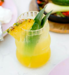 Tropical pineapple cocktail in a frosted curvy glass, layered yellow-green drink garnished with a pineapple wedge and green leaves on a white table in a bright brunch setting.