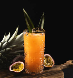 Tropical passion fruit cocktail in a tall textured glass on a wooden board, bright orange juice with ice, garnished with pineapple leaves and a dried citrus wheel, halved passion fruits and pineapple crown against a black background