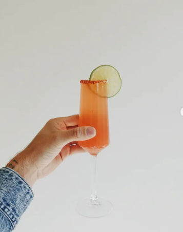 Denim-sleeved hand holding a champagne flute with a pink-orange cocktail, chili-salt rim and lime wheel garnish against a clean white background