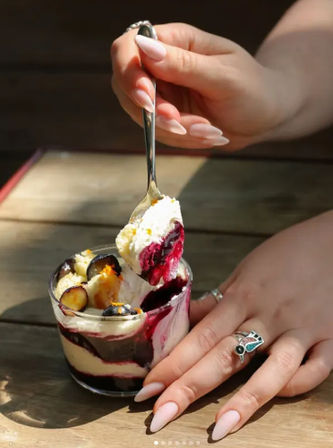 Hand with long pink almond nails and silver rings scooping a layered berry parfait in a small glass on a sunlit wooden table, creamy vanilla layers, dark berry compote and fresh blueberries.