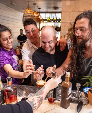 Group of friends at a cocktail bar enjoying a hands-on mixology moment, smiling as they stir a frothy shared drink with wooden swizzle sticks at a busy counter under warm pendant lights.