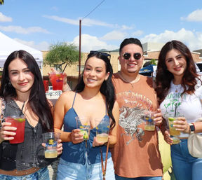 Four smiling adults holding colorful cocktails and cheersing at a sunny outdoor street festival with tents and vendor booths in the background.