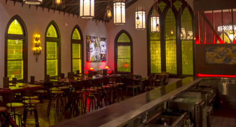 Cozy urban bar and restaurant interior with vaulted ceiling, Gothic arched stained-glass windows glowing green, long wooden tables and stools, hanging lanterns and warm red accent lighting along a polished bar counter.