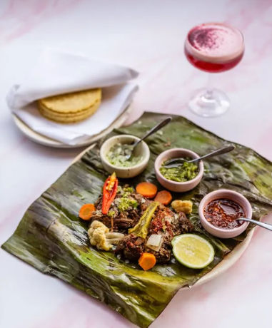 Vibrant taco plate: shredded meat with pickled vegetables and lime on a banana leaf, three salsas, stacked corn tortillas in a cloth and a pink cocktail