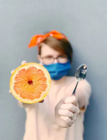 Halved grapefruit held toward the camera with a spoon, blurred person wearing a blue face mask and orange headband in the background