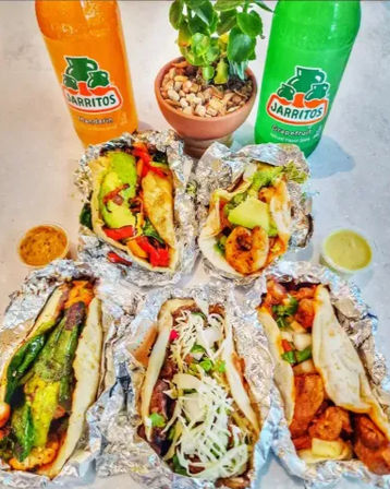 Six foil-wrapped Mexican street tacos filled with grilled vegetables, shrimp, avocado and cheese on a white table, with two colorful glass sodas and small salsa cups