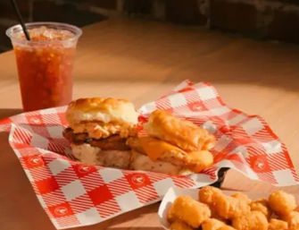 Southern-style breakfast: two biscuit sandwiches with egg, cheese and sausage on red-check paper, a side of crispy tater bites and a cold iced tea on a wooden table by a brick wall.