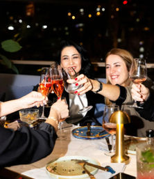 Cheerful group of friends toasting with rosé and wine glasses over a dinner table with empty plates and a small gold table lamp, city lights bokeh in the background.