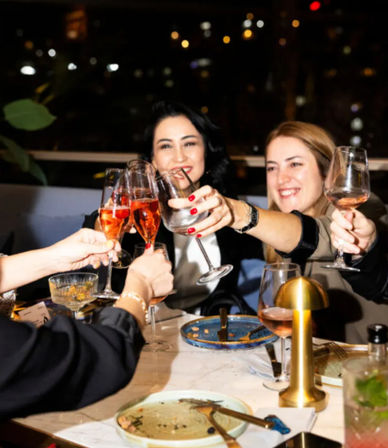 Cheerful group of friends toasting with rosé and wine glasses over a dinner table with empty plates and a small gold table lamp, city lights bokeh in the background.