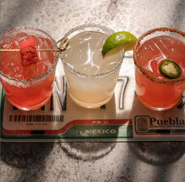 Mexico-themed flight tray with three colorful margaritas on a metal bar: a pink watermelon cocktail with a skewered cube, a pale lime margarita with a lime wedge, and a spicy jalapeño margarita with a chili-salt rim.