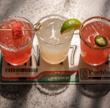 Mexico-themed flight tray with three colorful margaritas on a metal bar: a pink watermelon cocktail with a skewered cube, a pale lime margarita with a lime wedge, and a spicy jalapeño margarita with a chili-salt rim.