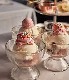 Three glass dessert bowls with vanilla ice cream scoops topped by small pink scoops and rainbow sprinkles on a white tablecloth — classic ice cream sundae display