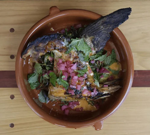 Charred whole grilled fish in a rustic clay bowl topped with pickled red onions, fresh herbs and spicy orange sauce on a wooden table