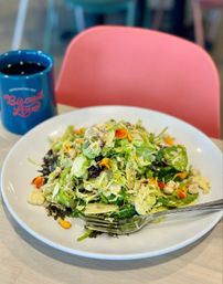 Shredded green salad with mixed greens, seeds and edible flower petals on a white plate, fork and blue coffee mug on a bright cafe table with a pink chair