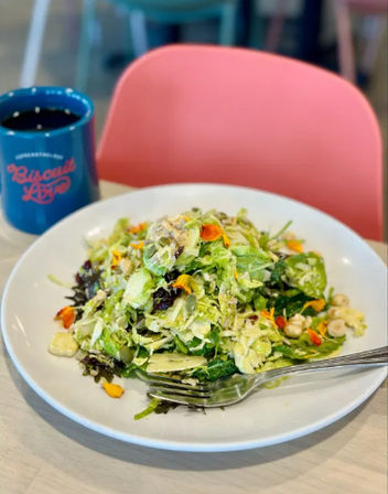 Shredded green salad with mixed greens, seeds and edible flower petals on a white plate, fork and blue coffee mug on a bright cafe table with a pink chair