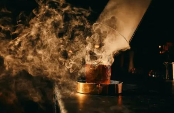 Smoked whiskey cocktail in a rocks glass on a brass coaster, amber drink with dramatic swirling smoke in a moody bar setting