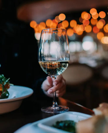 Hand holding a glass of white wine at a cozy dimly lit restaurant table with warm bokeh lights and plated dishes in the background