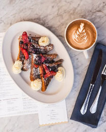 Berry-topped French toast triangles with whipped-cream rosettes and powdered sugar, served with a latte featuring leaf latte art on a marble café table