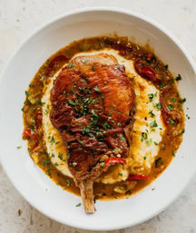 Seared bone-in pork chop on creamy mashed potatoes with a peppery pan sauce, red and green peppers, and chopped parsley garnish on a white plate — hearty restaurant-style comfort food.