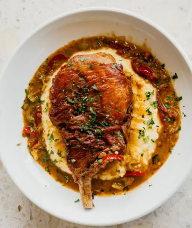 Seared bone-in pork chop on creamy mashed potatoes with a peppery pan sauce, red and green peppers, and chopped parsley garnish on a white plate — hearty restaurant-style comfort food.