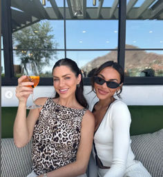Two friends smiling on a sunny rooftop patio lounge with a mountain view through large glass windows, one raising an orange cocktail for a celebratory brunch.